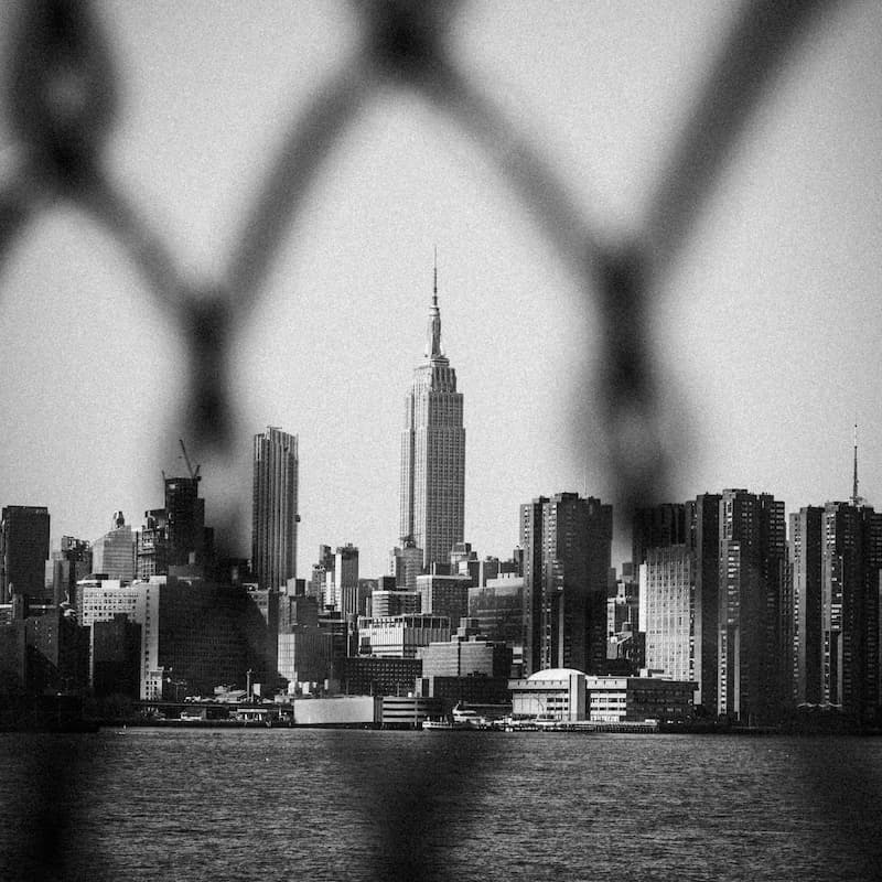 Someone looking through a chain link fence at New York City in black and white